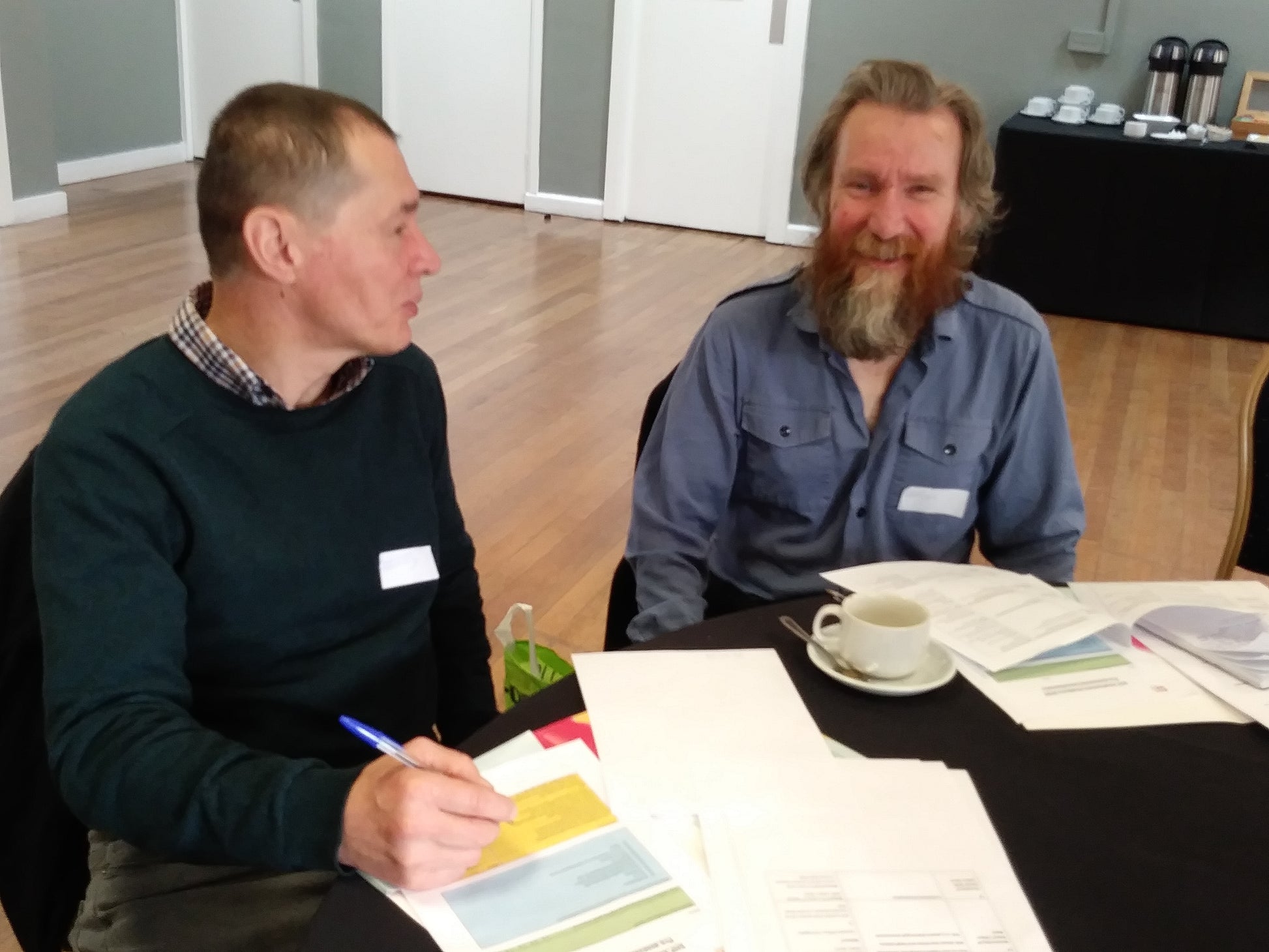 Two men sat around a table smiling with a cup of tea.