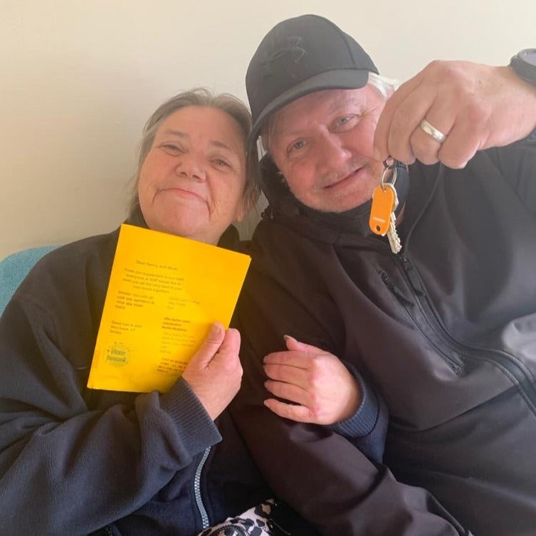 Woman smiling holding a yellow piece of paper sat next to a man smiling holding keys.